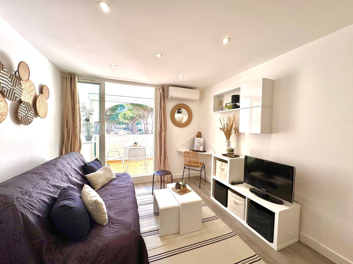 The living area features a spacious sofa with decorative cushions and a coffee table. A television is placed on a low white shelf. Natural light enters through large glass doors that open to a loggia with outdoor seating.
