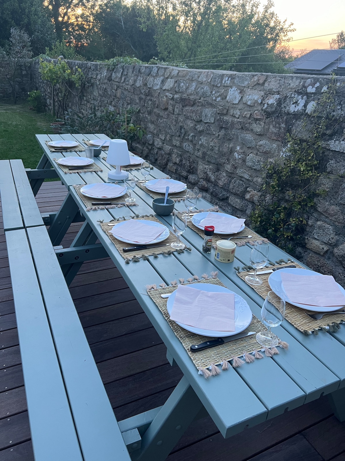 An outdoor dining table is set with plates, woven placemats, and glasses, inviting communal meals. The setting includes a tranquil garden area with a stone wall in the background, complemented by greenery, creating a peaceful atmosphere during the evening.