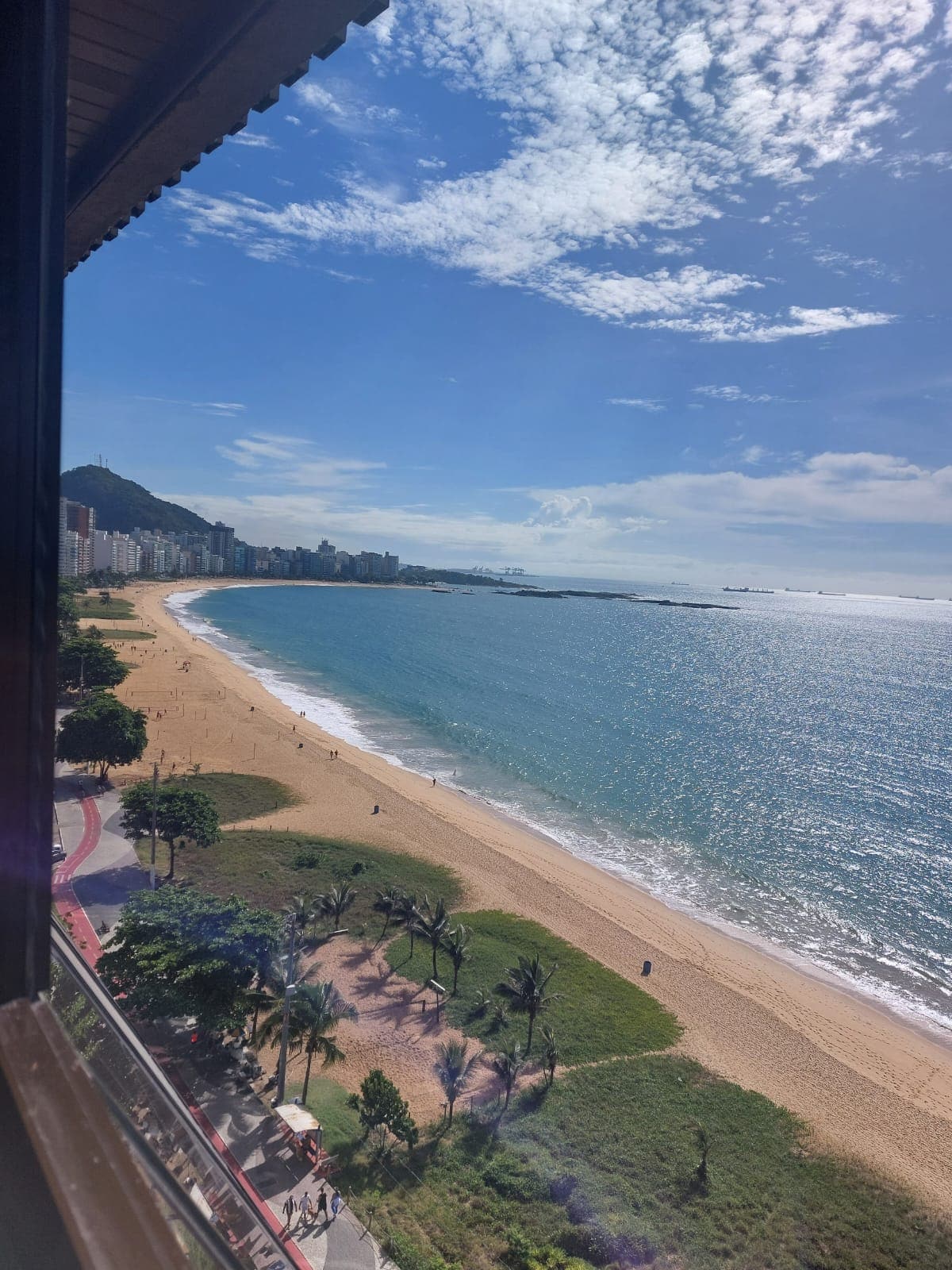 A scenic view showcases the golden sandy beach meeting tranquil blue waters under a cloudy sky. Palm trees line the beachfront, and a distant coastline is visible, providing a sense of spaciousness and calm. The scene reflects a peaceful coastal atmosphere.