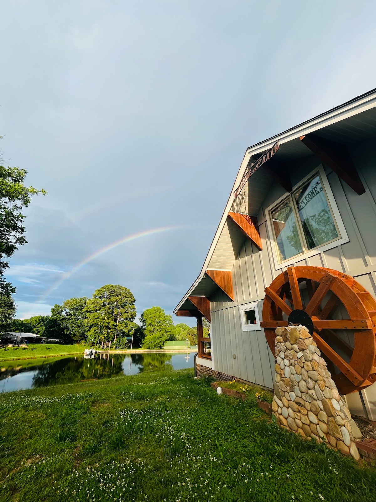 The exterior of the barn is highlighted by a wooden water wheel feature, with lush green grass and wildflowers surrounding the pond. A vibrant rainbow arcs across the sky, framing the scene and enhancing the natural beauty of the property.