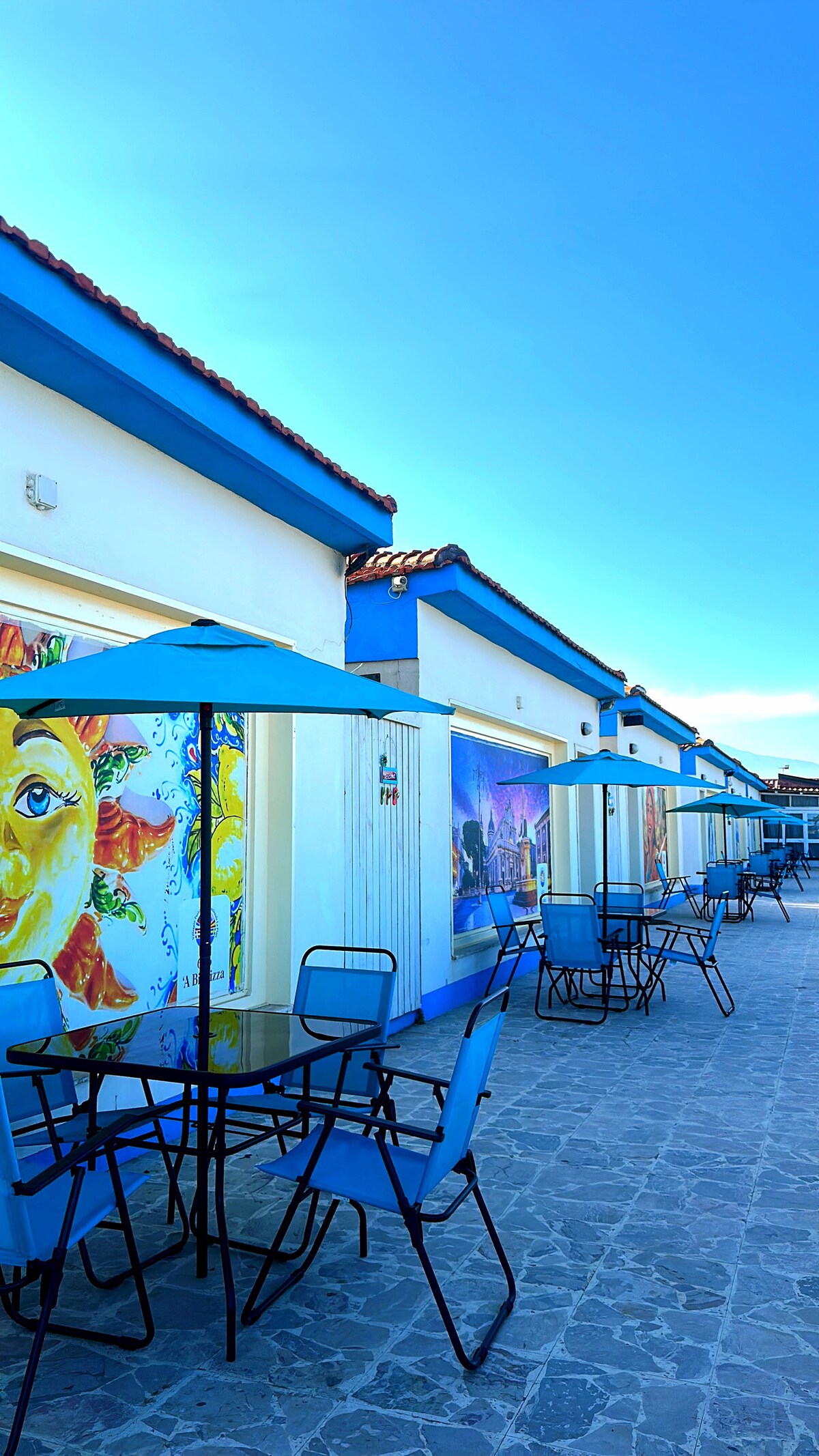 A row of colorful buildings is viewed against a clear blue sky. Each building features vibrant murals depicting sun and cultural themes. Outside, blue tables and chairs are placed under matching umbrellas on a stone-paved area, creating an inviting outdoor space.