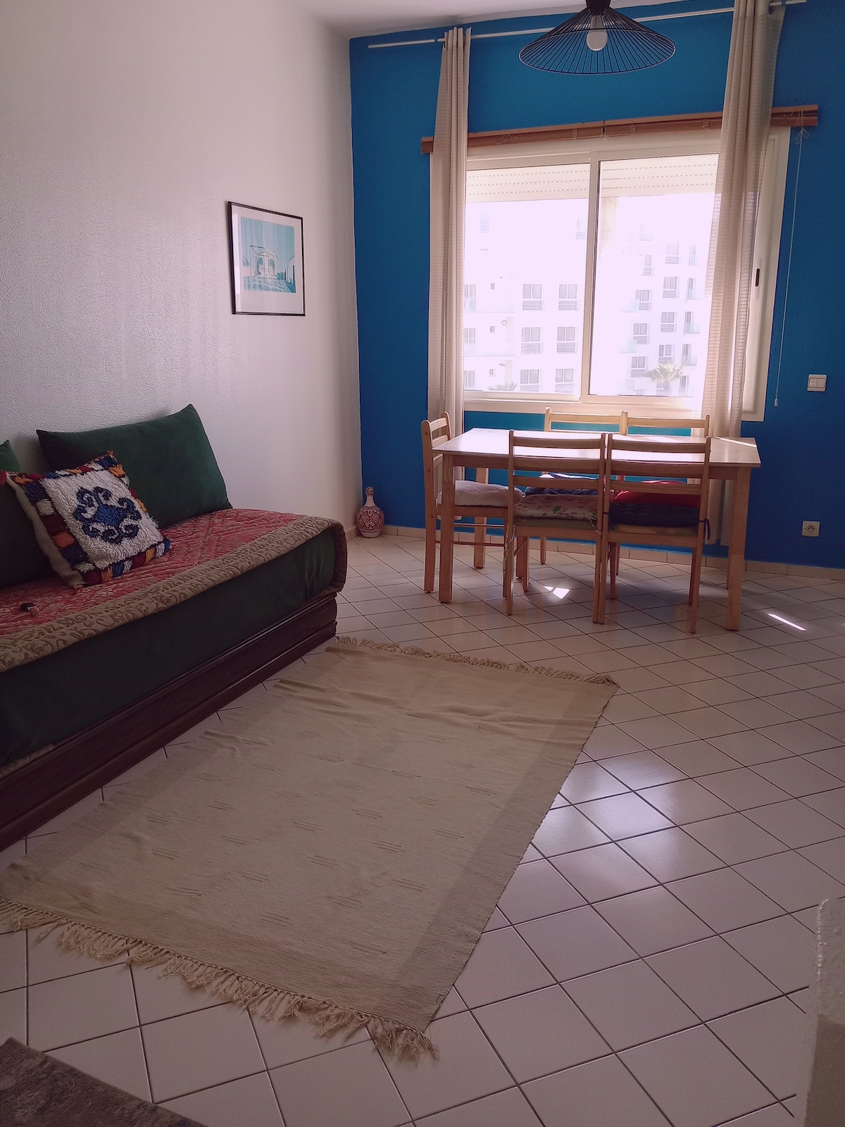 A bright living space features a cozy seating area with a green sofa and decorative pillows. A small dining table with four chairs is positioned near a window, allowing natural light to fill the room. A neutral rug lies on the tiled floor, contributing to the inviting ambiance.