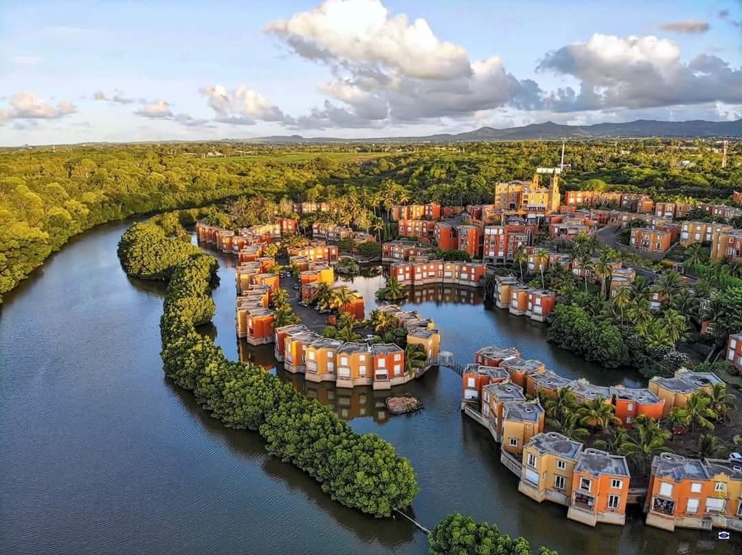 Aerial view of a coastal village features colorful buildings arranged along a winding river surrounded by lush greenery. The landscape creates a serene atmosphere with palm trees dotting the area, while the water reflects the colors of the structures, enhancing the picturesque environment.