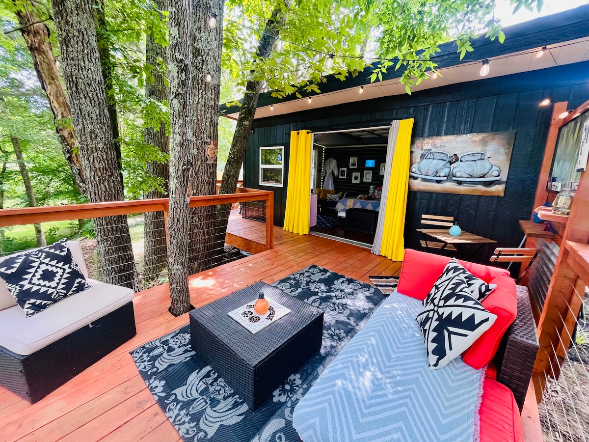 A deck area is surrounded by trees, featuring a red sofa and a patterned rug placed over wooden flooring. A coffee table sits in the center, while soft lighting extends from above. The open entrance to the cabin reveals a glimpse of the interior decor, with yellow curtains framing the entrance.