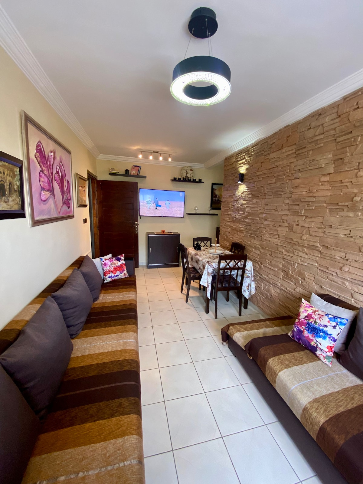 A cozy living space features two brown sofas adorned with colorful patterned cushions. A television is mounted on the wall, and a dining table is set for four, positioned near a decorative stone feature. Soft lighting from a modern fixture highlights the room's warm tones.