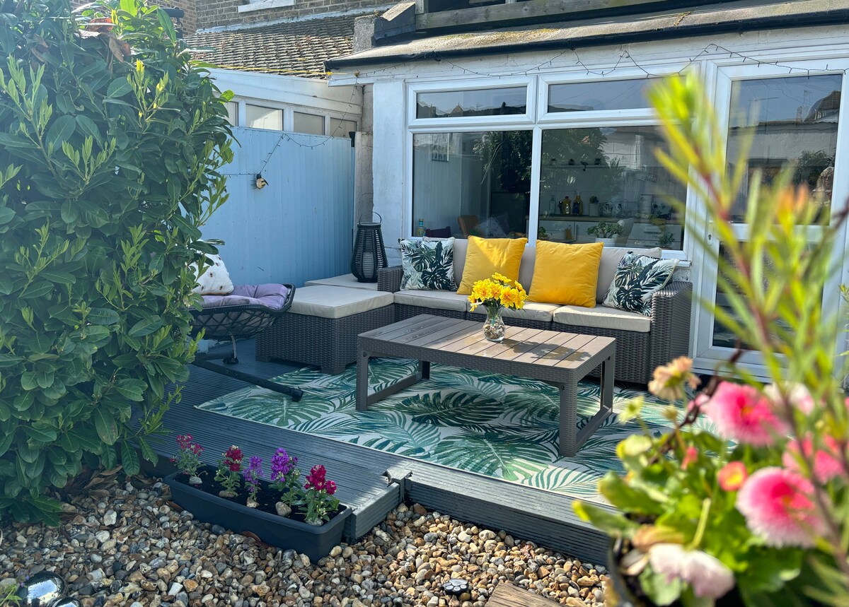 A well-furnished outdoor seating area features a comfortable gray couch adorned with yellow cushions. A coffee table is centered on a leaf-patterned rug, surrounded by vibrant flower beds. Lush greenery creates a sense of privacy, enhancing the inviting space for relaxation.