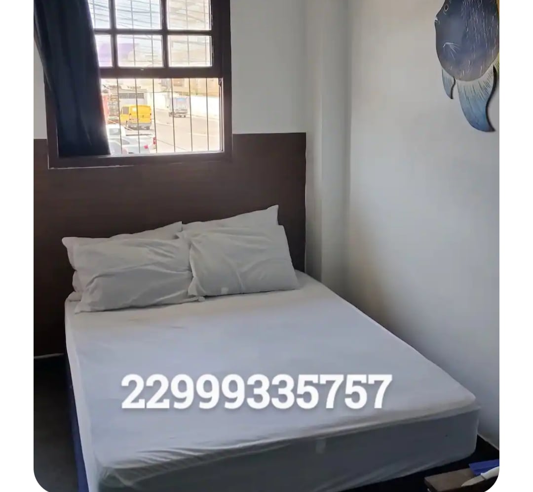 A cozy sleeping area features a spacious bed with four pillows arranged on top. Natural light enters through a window dressed in dark curtains, illuminating the room's simple design and a decorative wall piece of a fish.
