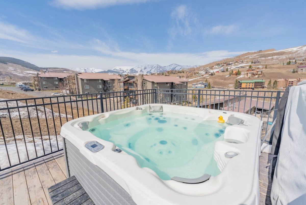 A hot tub is situated on a wooden deck, offering clear views of the surrounding mountains and nearby buildings. The water is invitingly calm, with a small rubber duck floating. The space is enclosed by a black railing, enhancing the open-air experience.