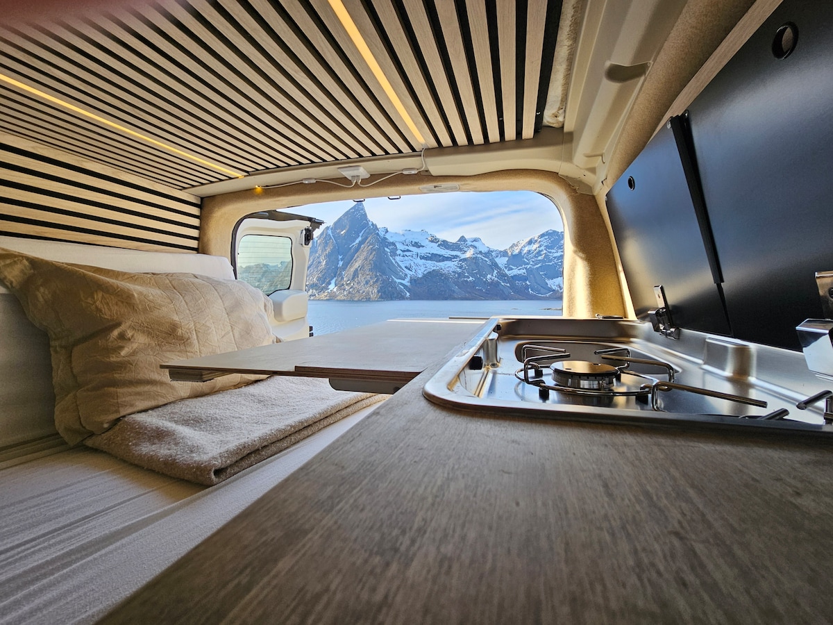 An interior view of the camper highlights a spacious bed and kitchen area. Visible are wooden plank ceilings and a compact cooking station with a gas burner. The large window frames a stunning mountain landscape, allowing natural light to fill the space.