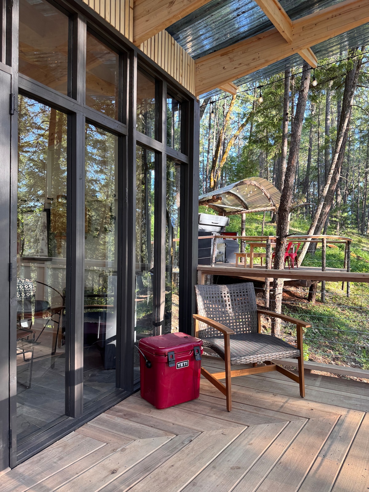 A modern cabin exterior features large glass doors that lead onto a wooden deck. A folding chair is positioned beside a Yeti cooler, with trees visible in the background. A second deck area is seen above, showcasing additional outdoor furniture.