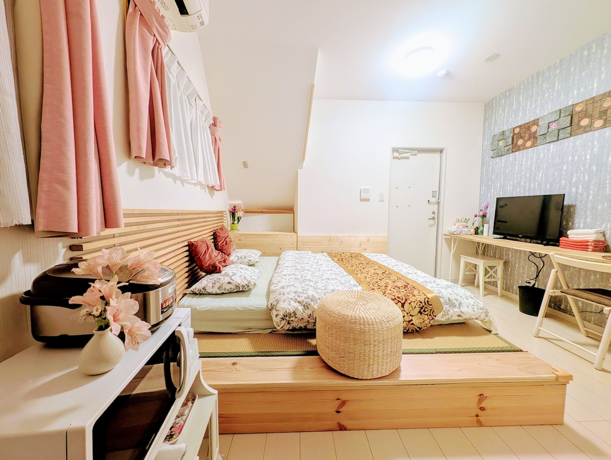 A spacious sleeping area features a large bed with decorative pillows, flanked by a wooden platform. Light-colored curtains frame the window, allowing natural light to illuminate the room. A small table and a TV are positioned across from the bed, offering essentials for relaxation.