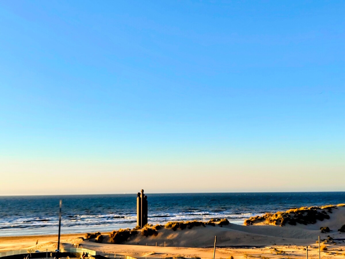 A serene view showcases the vast ocean meeting the sky at sunset, with soft sand dunes in the foreground. A structure is positioned along the shore, silhouetted against the horizon, enhancing the peaceful coastal landscape.