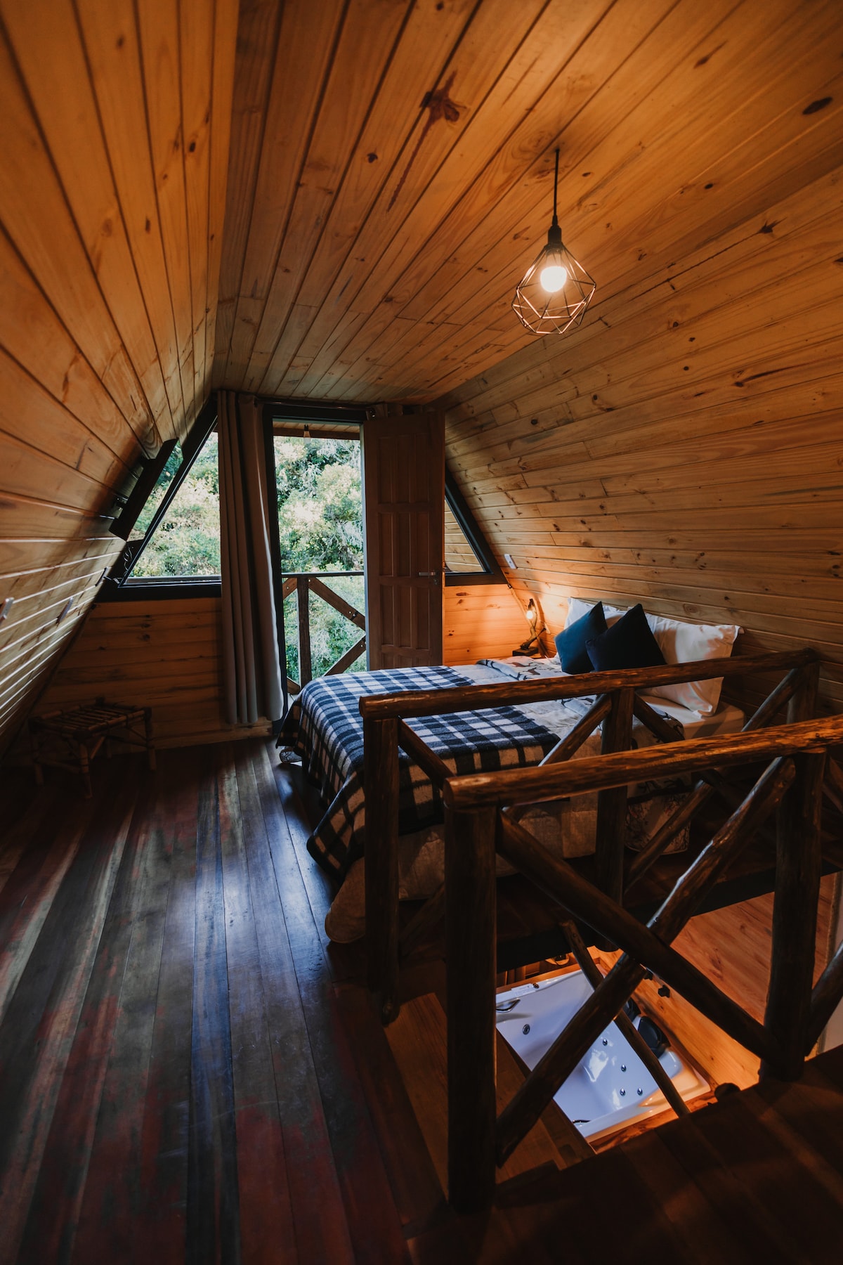 An inviting loft bedroom showcases slanted wooden walls and ceiling, contributing to a rustic feel. A cozy bed with a checkered blanket is positioned near triangular windows offering natural light and views of greenery. A warm light fixture adds a soft glow to the space.
