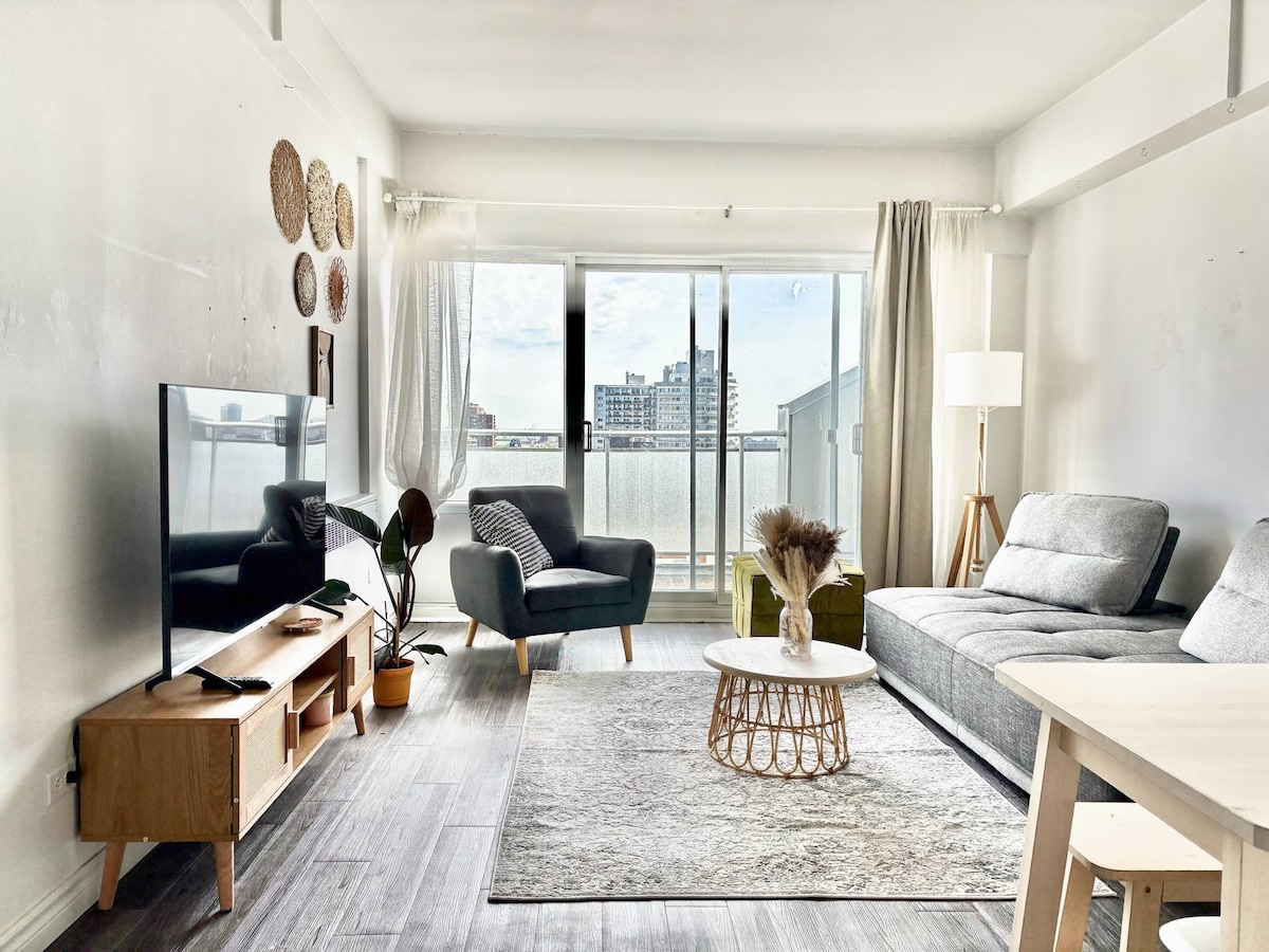 A bright living area is shown featuring a sectional sofa and an armchair, positioned around a coffee table. A large window provides natural light and a view of the city skyline. Decorative elements include wall art and a houseplant, contributing to a welcoming atmosphere.
