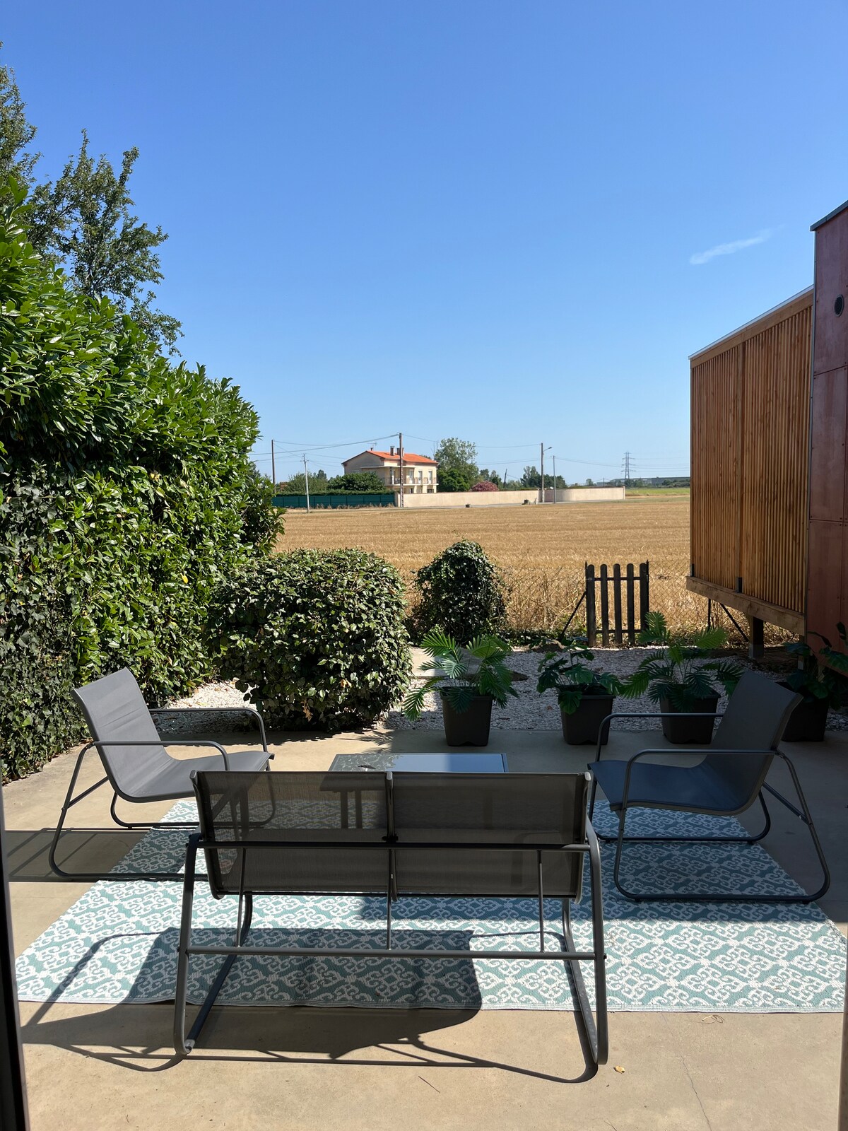 An outdoor seating area features a set of four chairs arranged around a small table, placed on a patterned rug. Lush green hedges and potted plants surround the space, with a view of open fields under a clear blue sky in the background.