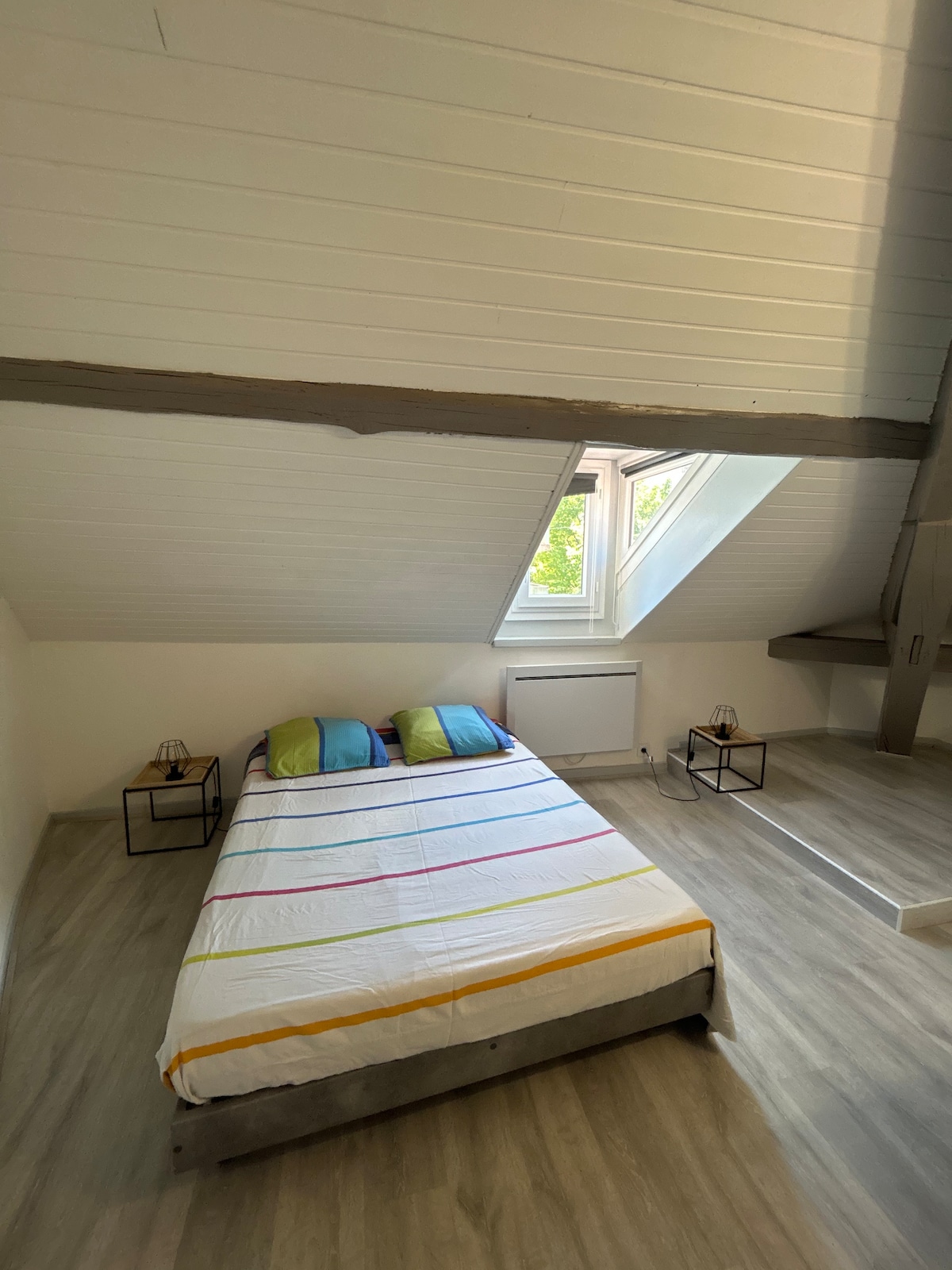 A cozy bedroom features a spacious bed with a striped blanket in tranquil colors. Two nightstands are placed beside the bed, each with a small lamp. A window with a slanted roof allows natural light to illuminate the room, highlighting the wooden beams overhead.