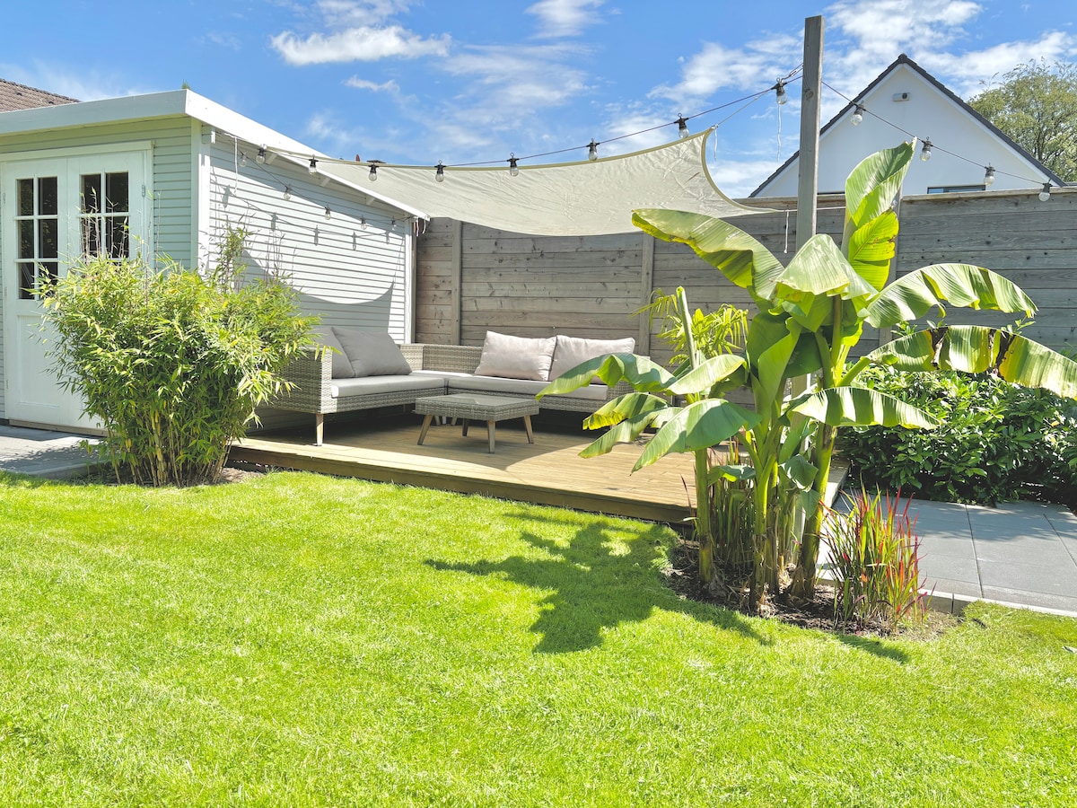 A garden space features a wooden deck with a light-colored seating area, shaded by a fabric canopy. Lush green plants border the area, and well-maintained grass enhances the inviting atmosphere. A glimpse of a small building is visible in the background, contributing to the garden's serene setting.