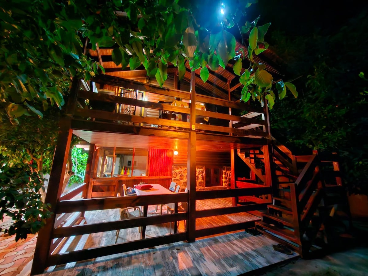 The wooden house is surrounded by lush greenery, highlighted by warm lighting at night. An outdoor dining area with a table and chairs is visible on the spacious deck, offering a welcoming space for gatherings. Stairs lead to the upper level of the residence.