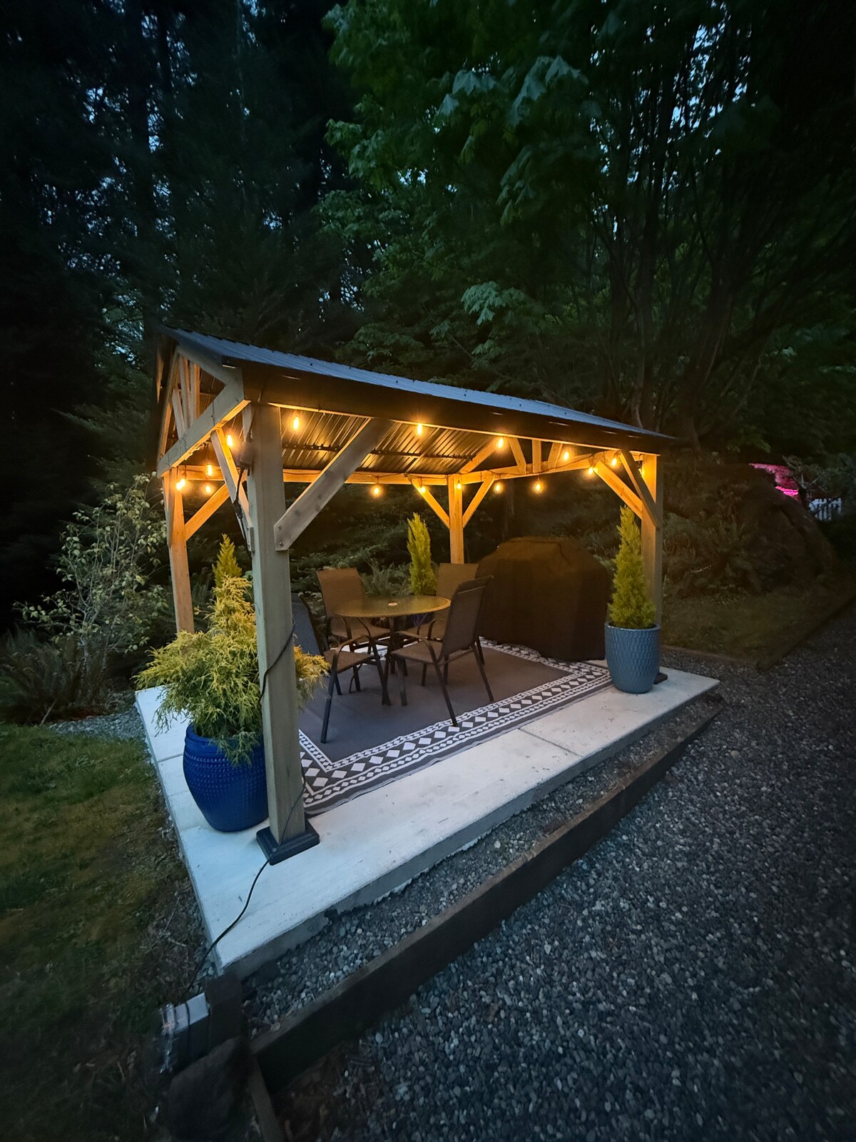 A gazebo is softly illuminated by string lights, creating a warm ambiance. Inside, a small table accommodates four chairs, surrounded by planters with greenery. The structure is set on a concrete pad, with a gravel path leading up to it, nestled among trees and plants.