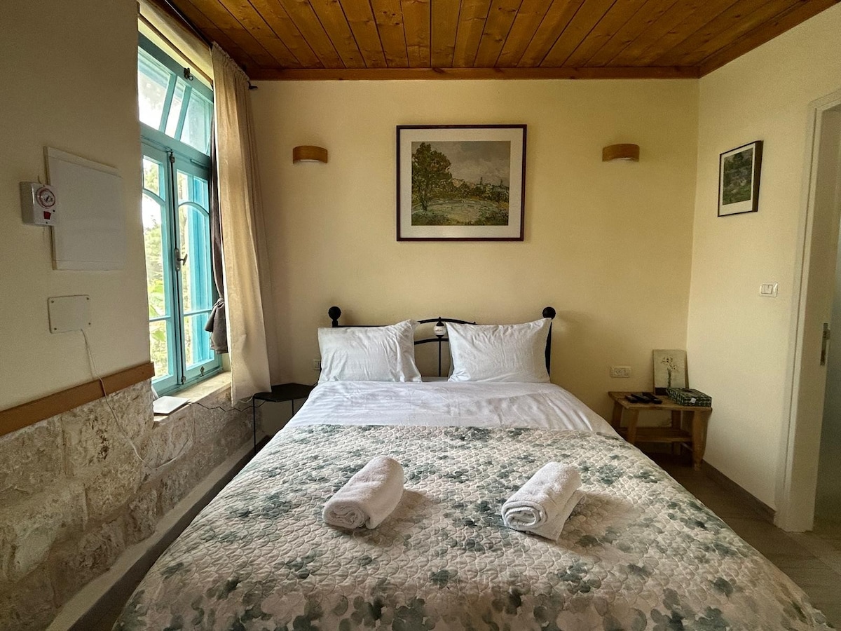 A well-appointed sleeping area features a spacious bed with a patterned quilt and fresh towels laid out. Natural light enters through a large blue-framed window, complemented by soft wall sconces and a framed landscape painting.
