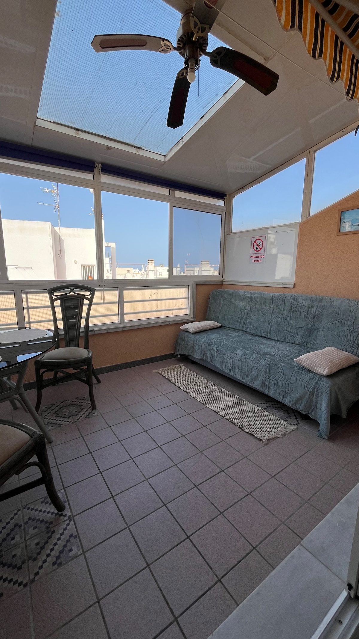 A spacious, enclosed terrace features large windows that invite natural light. A light blue sofa is placed against the wall, complemented by two small pillows. A round glass table with two chairs provides a cozy sitting area, while a woven rug adds texture to the tile floor.