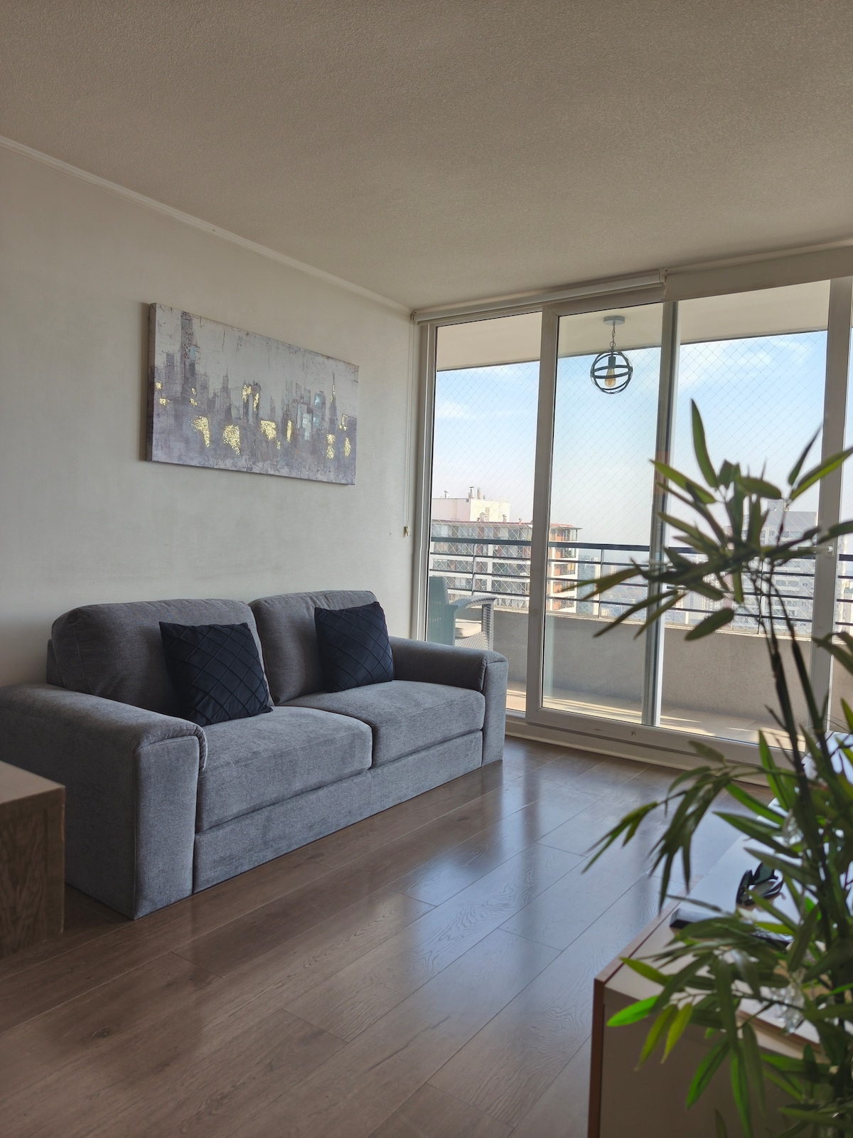 A gray sofa is positioned near a large window, allowing natural light to illuminate the space. A modern painting with subtle colors hangs above the sofa. A potted plant adds a touch of greenery to the area, enhancing the overall ambiance.