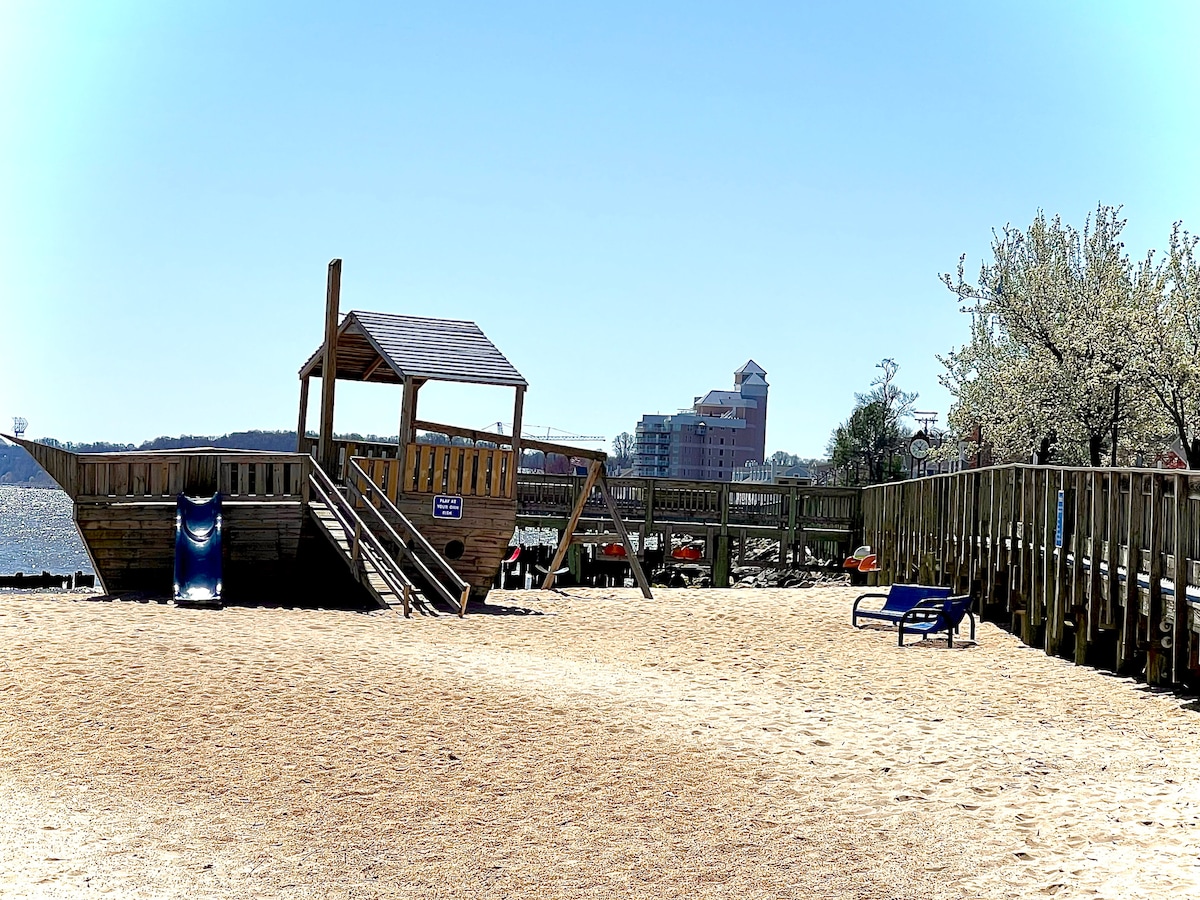 A sandy beach area is seen with a wooden play structure featuring a slide and stairs. Nearby, a blue lounge chair is positioned for relaxation. In the background, a multi-story building can be glimpsed along with trees lining the shore.