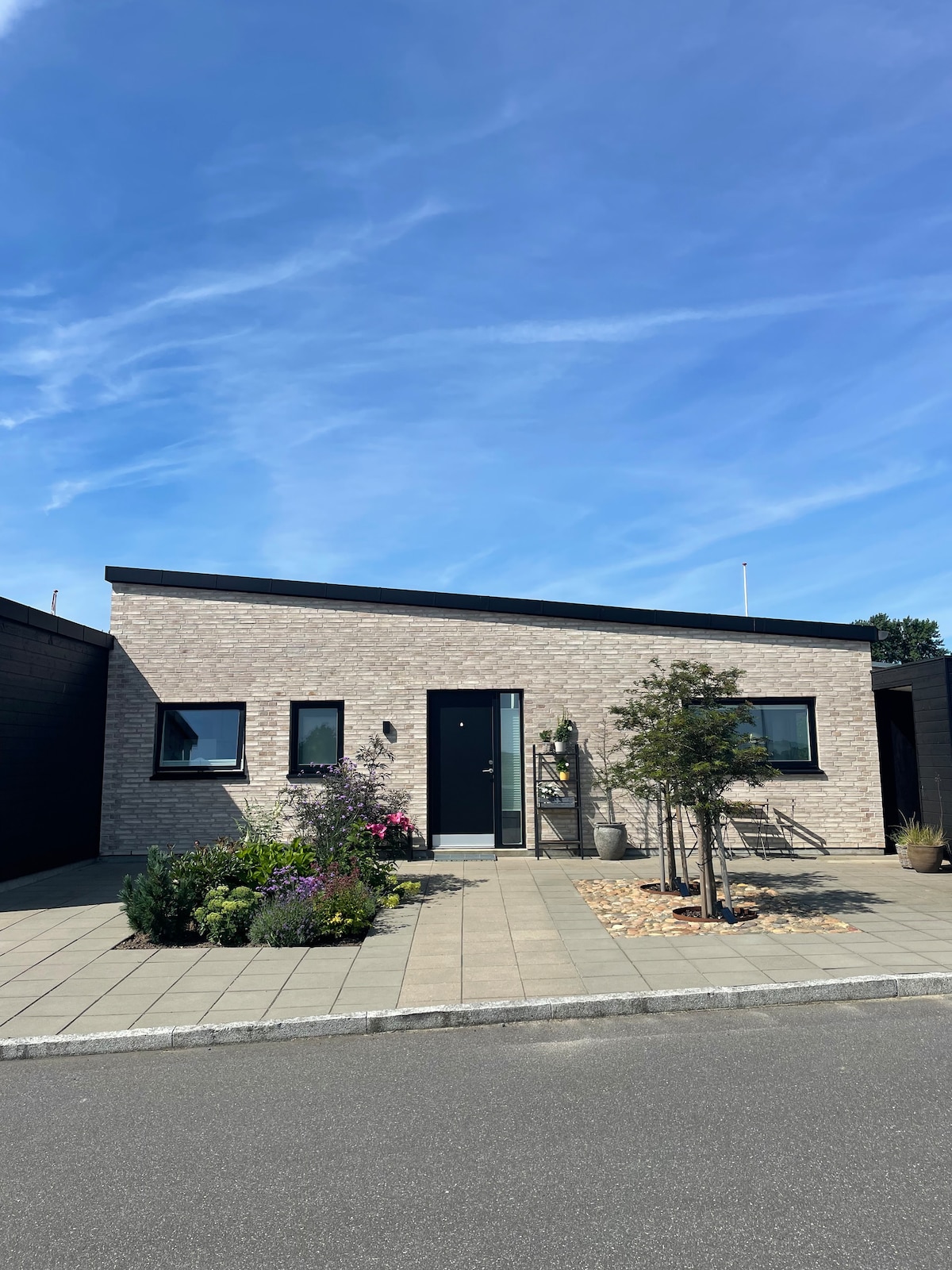 The modern exterior of the home features a light-colored brick facade with three large windows, framed by a well-maintained garden of colorful flowers. A clear blue sky can be seen above, enhancing the inviting appearance of the entrance.