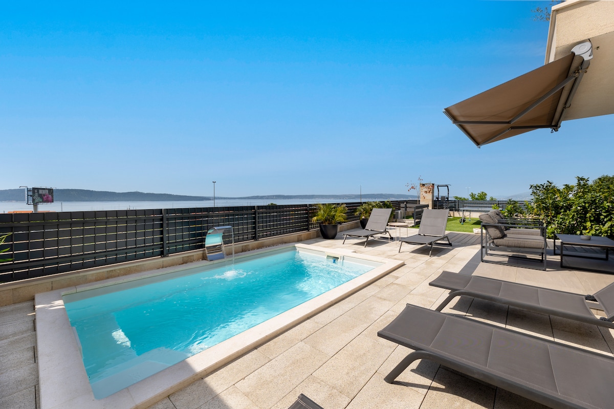 A private heated swimming pool is surrounded by a spacious deck, featuring lounge chairs for relaxation. The area is bordered by potted greenery and a sleek fence, with views of distant hills and the coastline, creating a serene outdoor setting.