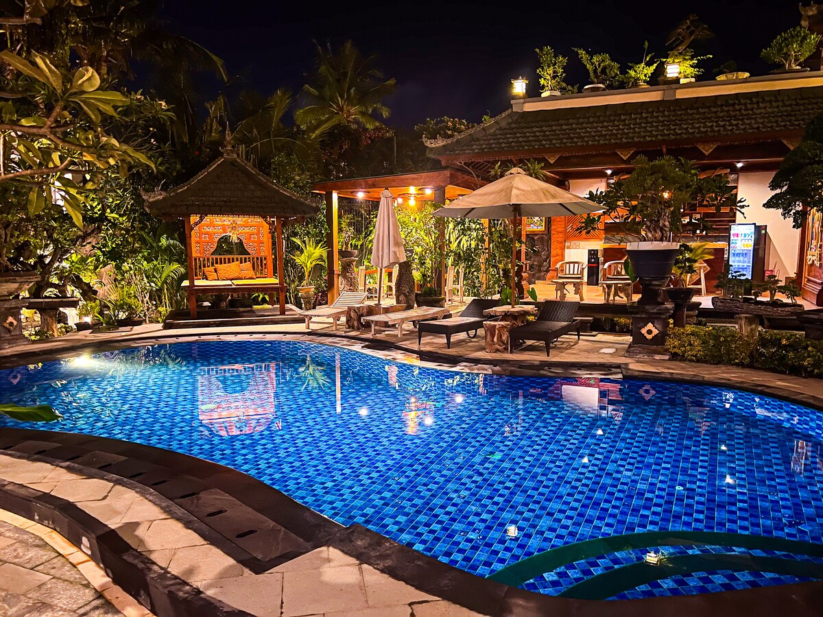 A tranquil nighttime scene features a clear blue pool surrounded by lush greenery and tropical plants. Multiple lounge chairs are arranged nearby, alongside a shaded seating area with a wooden gazebo. Soft lighting enhances the inviting atmosphere of the space.