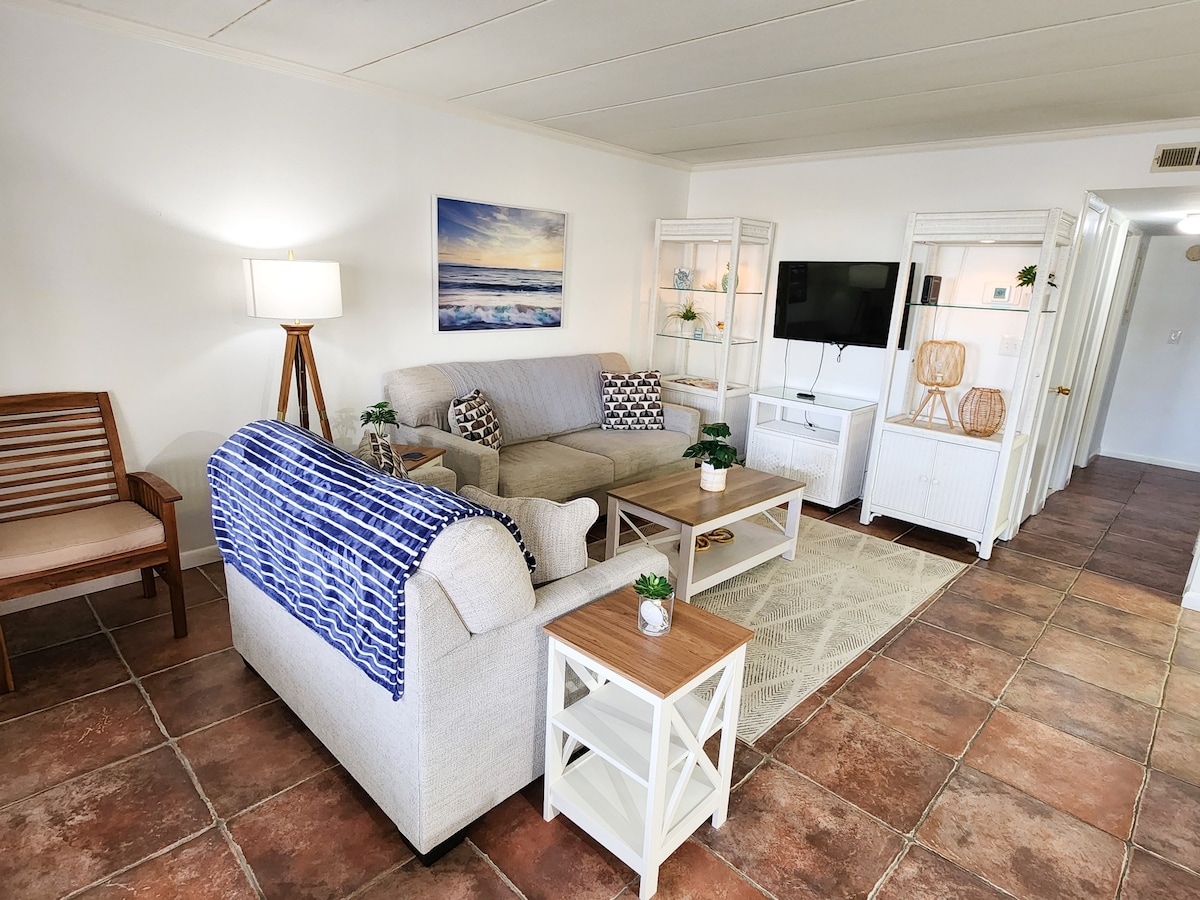 A welcoming living area features a light-colored sofa and armchair, complemented by textured throw pillows. A flat-screen television is mounted on the wall, while a small coffee table sits in the center. Decorative shelving displays items, and soft lighting enhances the space.