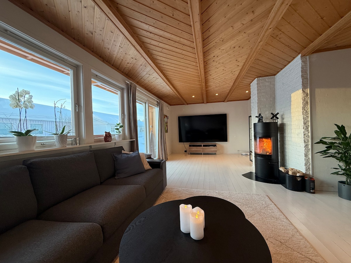 A spacious living area features a modern dark sofa arranged around a circular coffee table with lit candles. A large television is mounted on a white wall, and a wood-burning stove adds warmth. Natural light fills the room through multiple windows, highlighting the wooden ceiling.