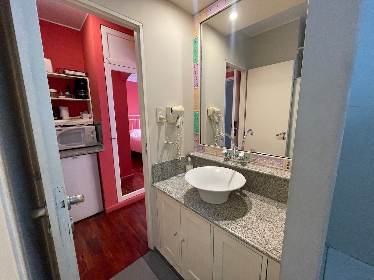A compact bathroom is presented featuring a modern sink with a round basin set upon a granite countertop. Mirrors on the walls reflect the bright light. A small kitchenette is visible in the background, along with an entryway leading to a vibrant red-painted room.