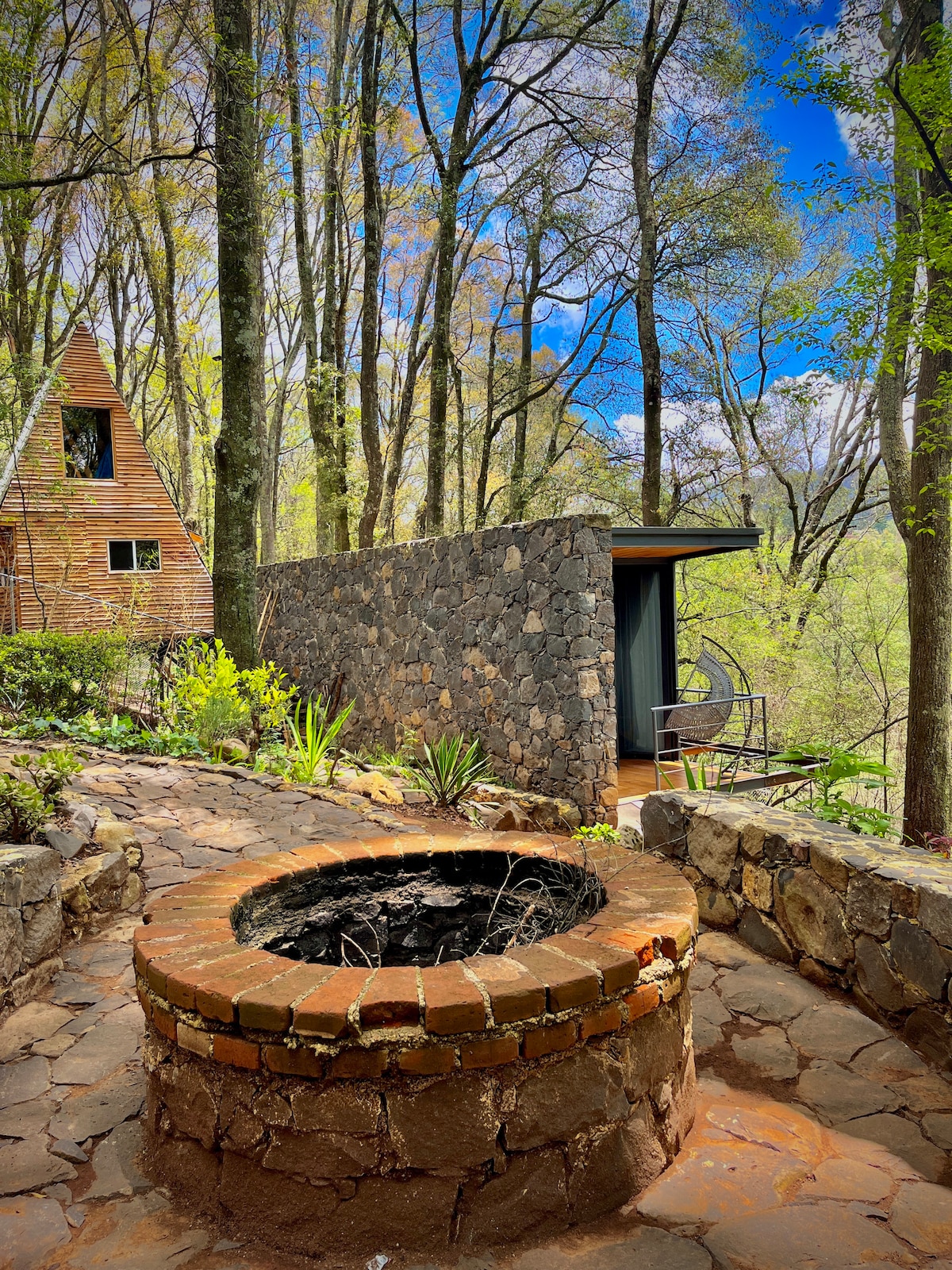A stone pathway leads to a circular brick fire pit, surrounded by lush greenery and trees. The backdrop features a modern A-frame structure and a stone wall, creating a natural setting that harmonizes with the environment.