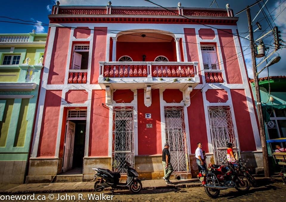 The exterior of a vibrant pink building showcases its original architectural details, including intricate white wrought ironwork on the windows and doors. A balcony extends above the entrance, adding character to the structure. Nearby, motorcycles are parked along the cobblestone street.