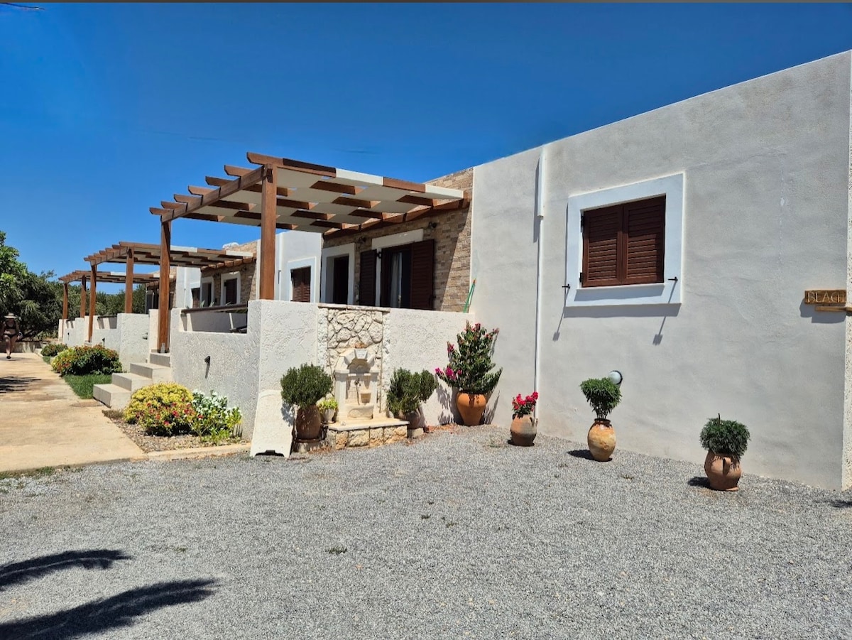 The exterior of the Olivar Suites is depicted with stone and whitewashed walls, complemented by wooden pergolas. Potted plants and flowers are arranged along the walkway, offering a serene entryway. A gravel driveway leads to the suites, surrounded by green foliage.