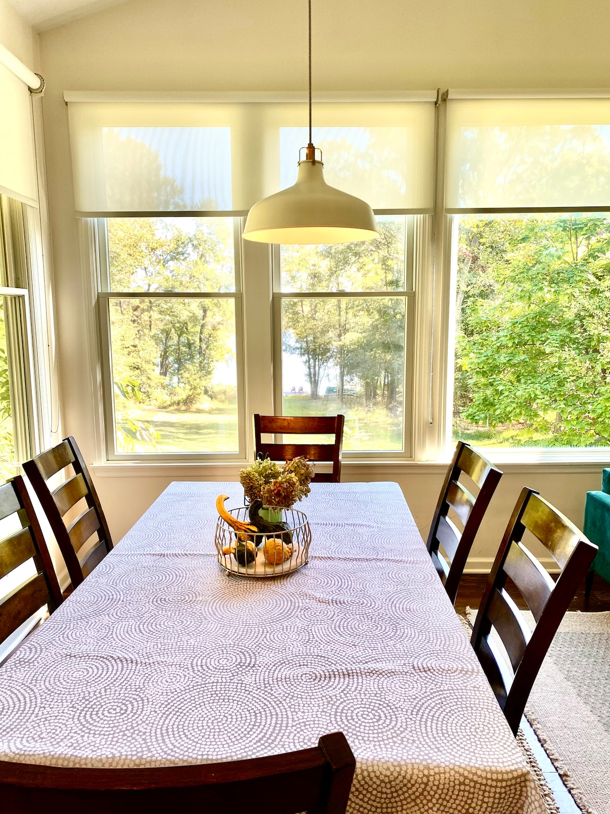 A dining table seating six is positioned in a bright room with large windows showcasing views of the surrounding trees and the lake. A simple table runner centers a basket containing fruits, while soft natural light enhances the inviting atmosphere.