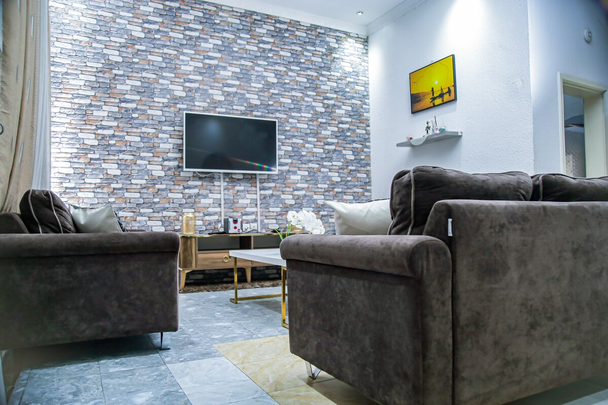 A spacious living area features a stylish stone accent wall, complemented by two plush sofas and a modern coffee table. A flat-screen television is mounted on the wall, and soft lighting enhances the inviting ambiance of the space.
