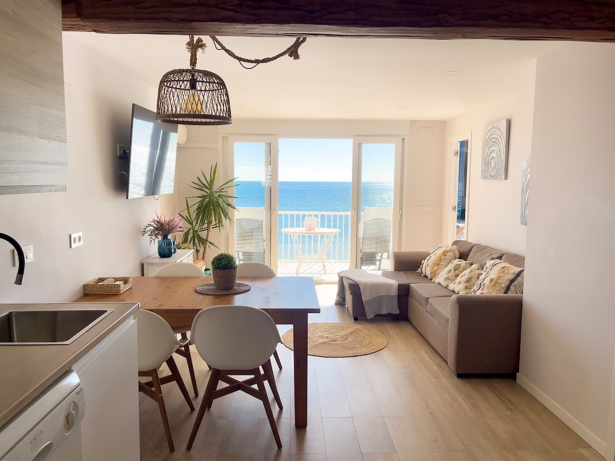 An open-plan living area showcases a comfortable sofa and a dining table with white chairs, all set against a backdrop of large glass doors. The view features the blue ocean and picturesque balcony, with indoor plants adding a touch of greenery.