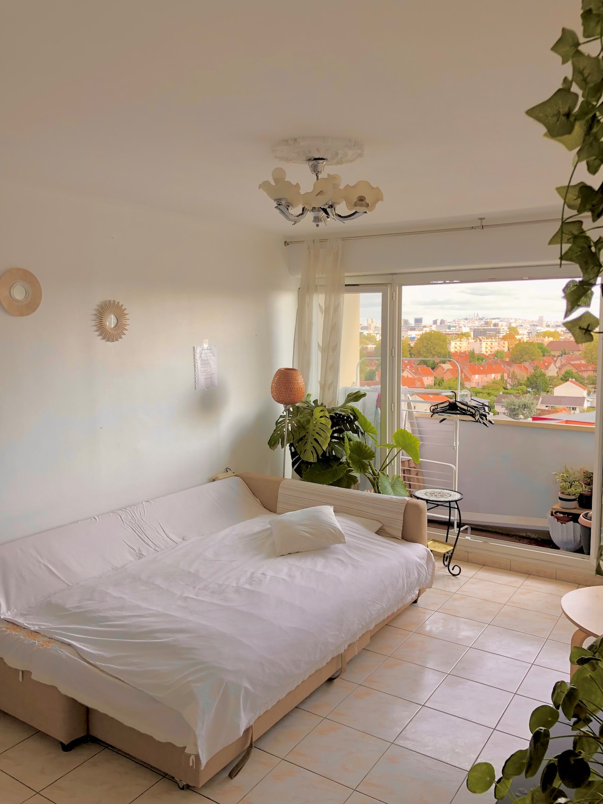 A bright living area is furnished with a sofa bed, complemented by a white blanket and cushion. A large window offers city views, while potted plants add greenery to the space. Simple decorative elements, including a ceiling light fixture and wall hangings, are present.