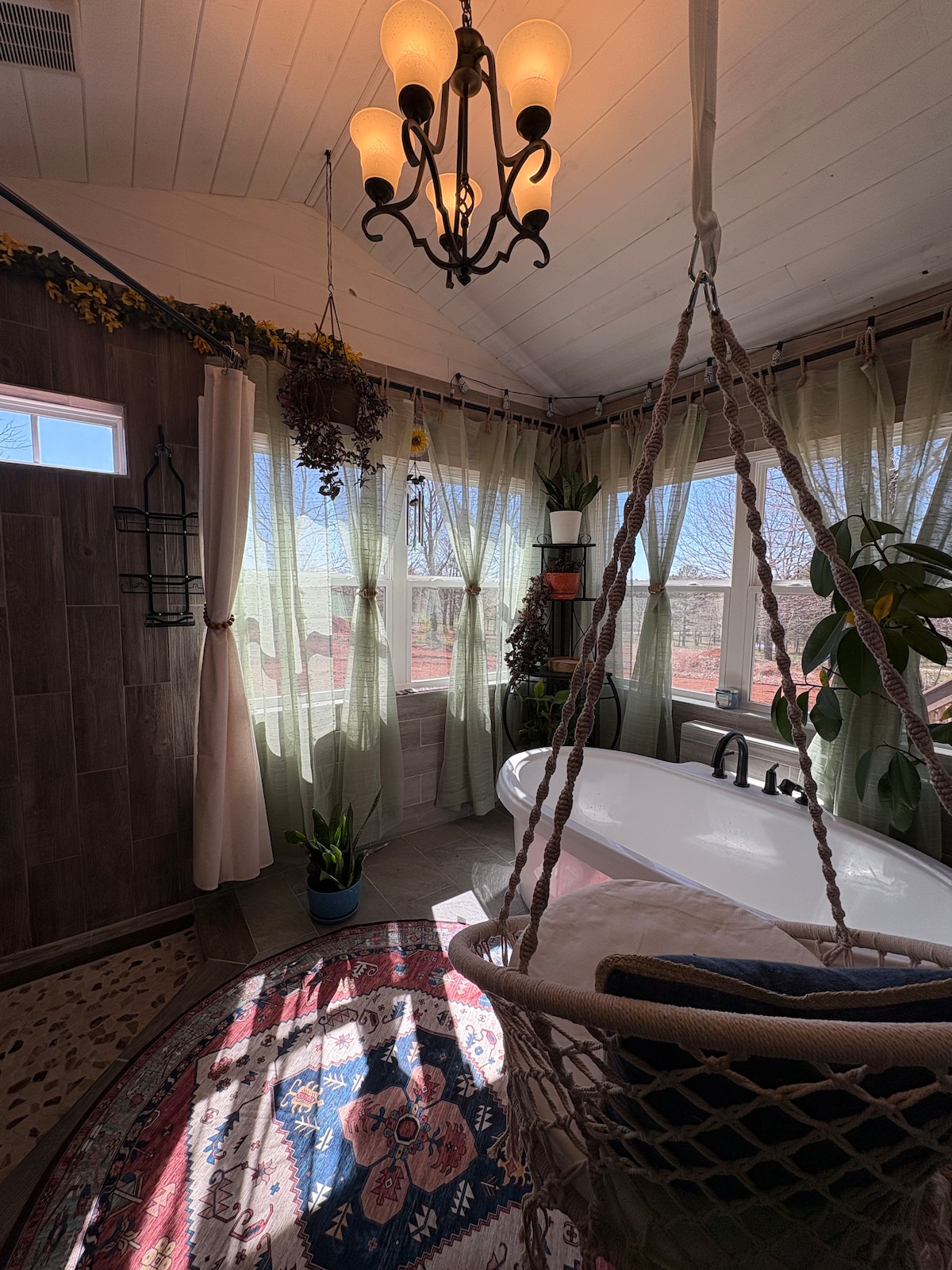 A cozy bathroom space features large windows with soft green curtains allowing natural light to filter in. A hanging boho chair offers a relaxing spot, while a freestanding bathtub invites restful soaks. Plants adorn the area, enhancing the tranquil ambiance. A patterned rug adds warmth underfoot.