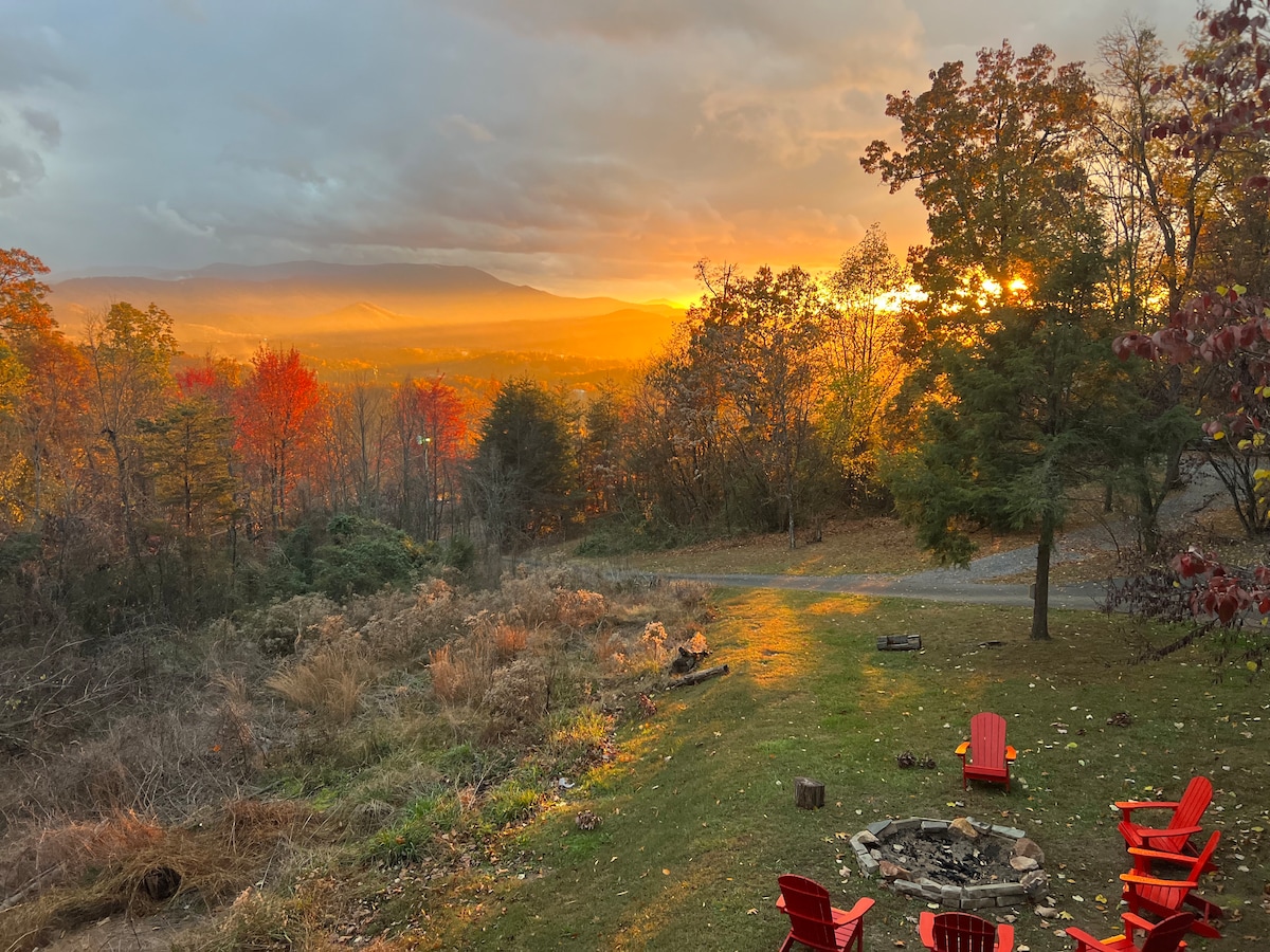 An outdoor seating area features several red chairs arranged around a fire pit, set against a backdrop of colorful autumn trees. In the distance, mountains are visible under a dramatic sky, with warm hues of orange and yellow illuminating the scene as the sun sets.