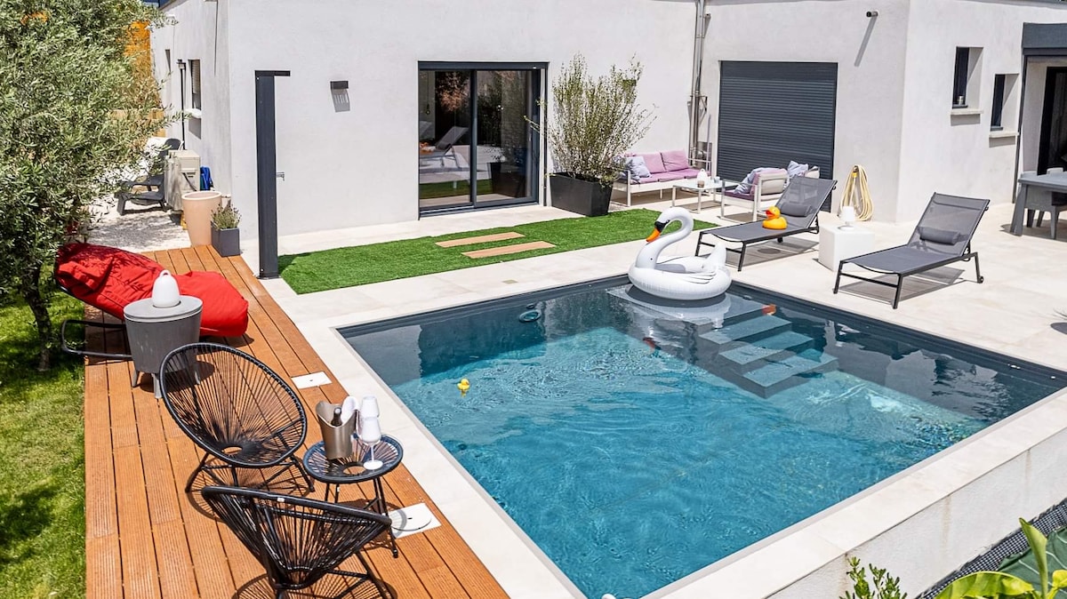 An outdoor area features a refreshing pool surrounded by lounge chairs and a wooden deck. A large inflatable swan floats in the water, and colorful toys are also visible. The space is complemented by green grass and decorative plants, creating an inviting environment.