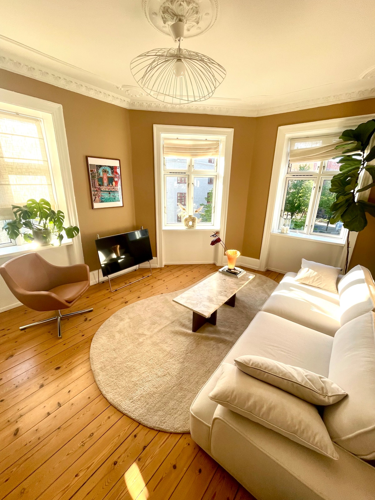 A well-lit living area features large windows allowing natural light to fill the space. A round rug lies beneath a wooden coffee table, with a comfortable sofa and a stylish chair positioned nearby. Potted plants add greenery to the room's warm tones.