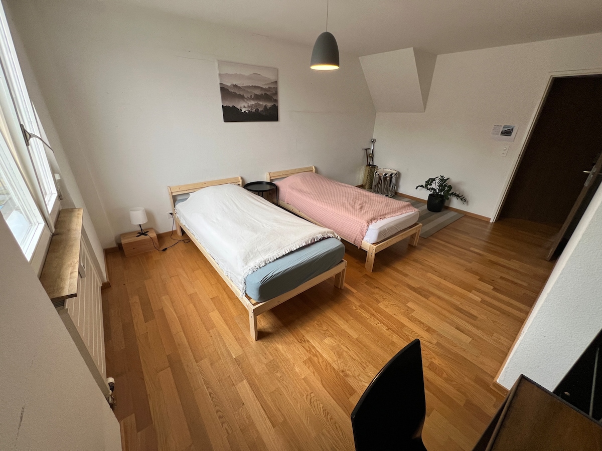 The sleeping area features two spacious beds placed parallel to each other, each adorned with simple bedding. Natural light streams in through a large window, highlighting the warm wooden floor. A small bedside lamp and a black chair are visible, contributing to the functional layout.