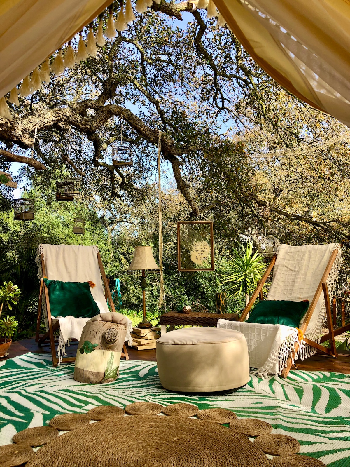 A serene seating area is featured under a canopy, with two reclining chairs equipped with soft green cushions. A small table is positioned nearby, accompanied by a cozy round ottoman and decorative plants. A textured rug with a leaf pattern enhances the relaxed atmosphere.