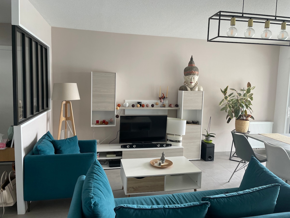 The living area is arranged with a turquoise sofa facing a modern entertainment center. A contemporary lamp stands beside the sofa. Light-colored furniture complements the space, which is enriched by a potted plant, creating a welcoming environment. A dining table is positioned nearby.