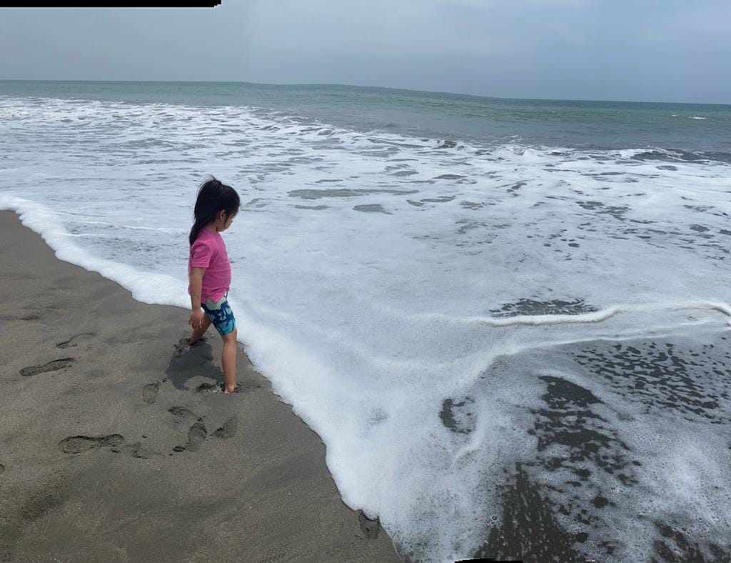 A child stands at the shoreline, where the gentle waves meet the sandy beach. The water appears frothy as it curls around the child's feet. The background reveals a vast ocean under a cloudy sky, creating a serene coastal scene.