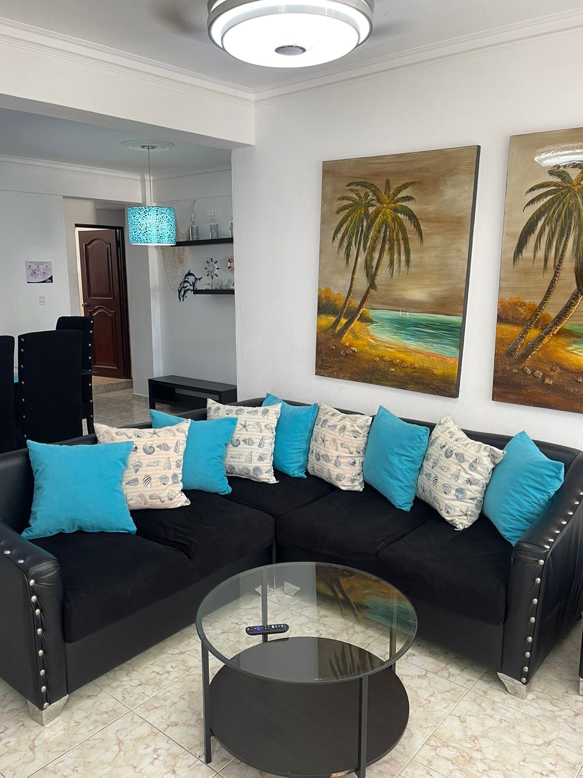A modern living area features a black sectional sofa adorned with blue and patterned cushions. A round glass coffee table sits in the center. Two framed palm tree artworks decorate the walls, complementing the natural light from nearby sources.
