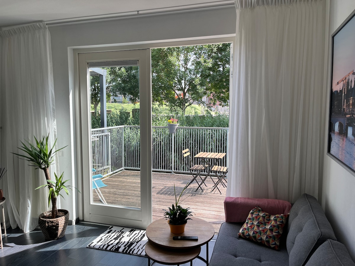 A bright living area features sliding glass doors that open to a spacious balcony, revealing greenery outside. A small outdoor table and chairs are visible on the deck, complemented by a cozy seating area with a coffee table and potted plant.
